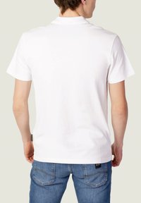 T-shirt en coton blanc avec une coupe classique, des manches courtes et une texture subtile. Présente une petite étiquette de logo près de l'ourlet.