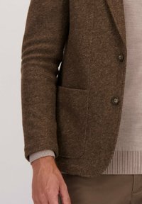 Man in een bruin wollen blazer over een lichtbeige trui en een bruine broek, met knopen en een voorzak op de blazer.