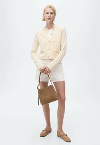Cárdigan de textura cremosa con cuello redondo y botones, combinado con una falda mini blanca. Sosteniendo un bolso de ante marrón y usando zapatos beige.