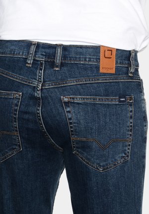 Blue denim jeans featuring a tan leather tag at the waistband, two back pockets with a distinctive stitching pattern, and a fitted design.