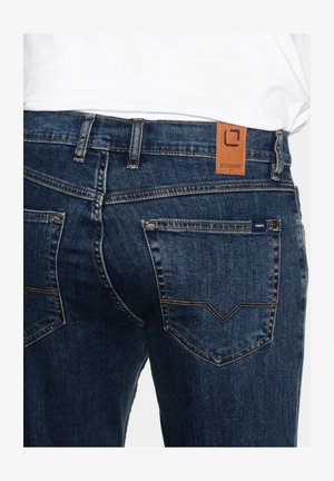 Blue denim jeans featuring a tan leather tag at the waistband, two back pockets with a distinctive stitching pattern, and a fitted design.