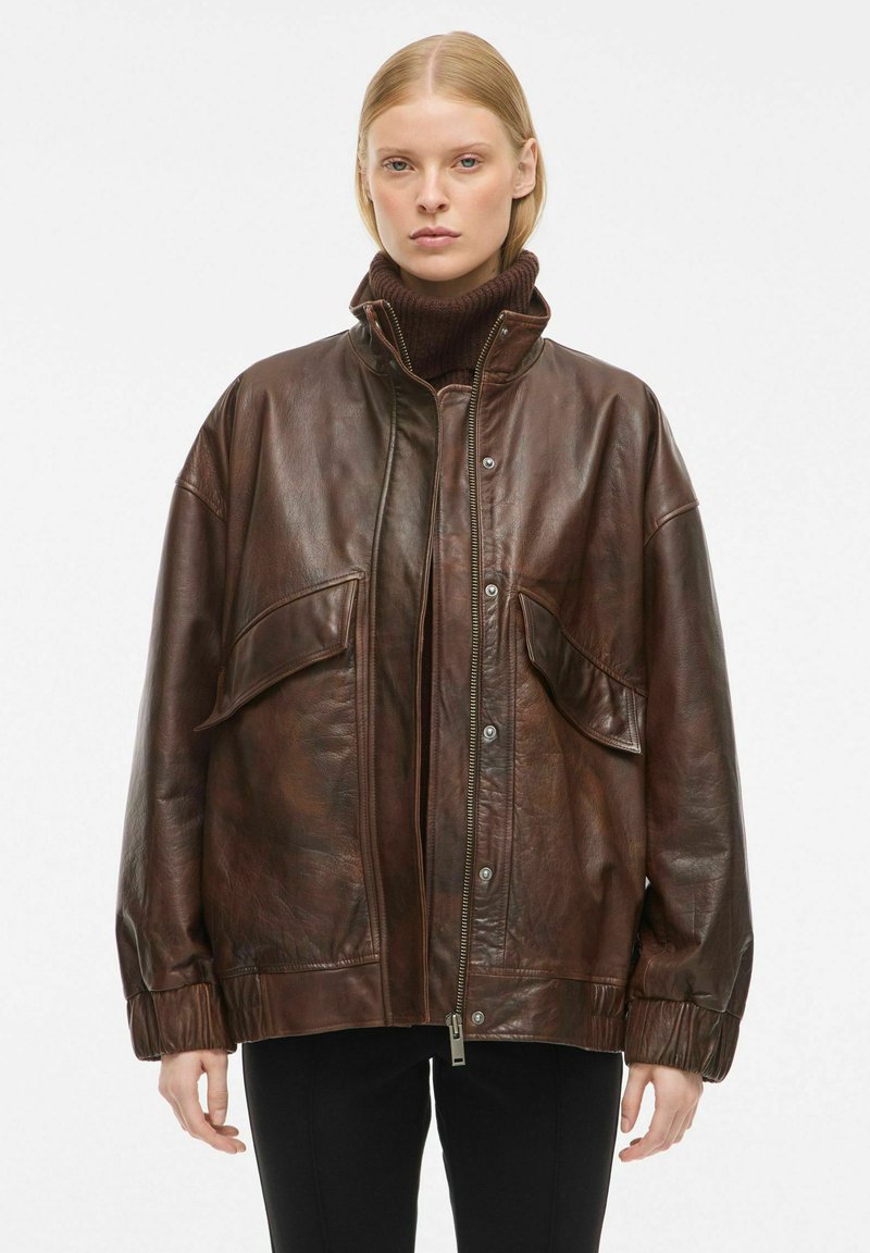 Pruunist nahast jakk, millel on lühendatud disain, kaks esikotti, tõmblukk ja soonikkoes mansetid ja krae. Sile pind.