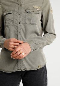 Zielona, długorękawowa koszula zapinana na guziki z dwoma kieszeniami na piersi, metalowymi guzikami i naszywką z logo na dole. Tkanina o lekko fakturowanej powierzchni.