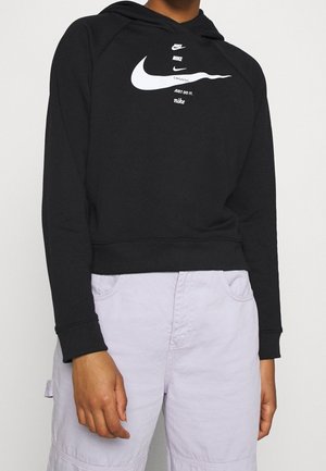 Osoba ubrana w czarną bluzę Nike z kapturem z wieloma logo i napisami, do tego jasnofioletowe spodnie na jednolitym tle.