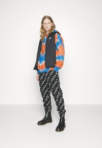 Crni sansruku prsluk preko kapuljače s višebojnim tie-dye uzorkom (narančasta i plava). Crne hlače s bijelim natpisom "JORDAN". Crne čizme do gležnja.