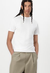 White short-sleeve t-shirt made of soft fabric, paired with light beige trousers featuring pleats and a relaxed fit. Model stands hands in pockets.