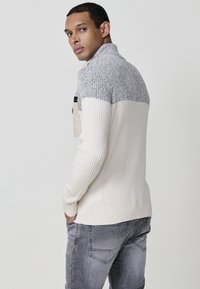 Suéter acanalado en crema y gris con un collar alto y hombros de punto gris, que cuenta con bolsillos laterales y un diseño ajustado.