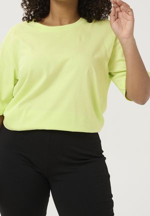 Woman with curly hair wears a loose light green shirt and fitted black pants, lifting one arm with fingers slightly bent.