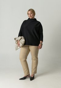 Zwart oversized top met een hoge kraag, beige nauwsluitende broek en zwarte slingback-hakken. Houdt een metalen zilveren tas vast met een getextureerd oppervlak.