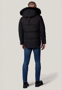 Czarna kurtka puchowa z kapturem wykończonym futrem, o dłuższym kroju, z zamykanymi bocznymi kieszeniami i gładką teksturą. Noszona z niebieskimi dżinsami i czarnymi botkami.
