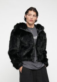 Casaco de pelo sintético preto com capuz, apresentando um fecho de zip e detalhes em botão, combinado com uma t-shirt cinza e calças pretas com padrão.