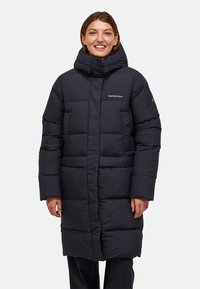 Leende person klädd i en lång svart vadderad vinterrock med hög krage och sidofickor, stående mot en vit bakgrund.
