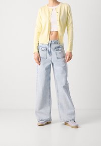 Geel cropped vest, witte crop top, lichtblauwe brede broek met zakken aan de voorkant en beige instap schoenen. Een zachte textuur overal.