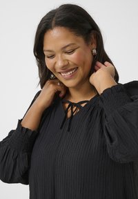 Blusa nera con texture, maniche a palloncino e dettagli con cintura al collo. Caratterizzata da un materiale leggero con un sottile motivo increspato.