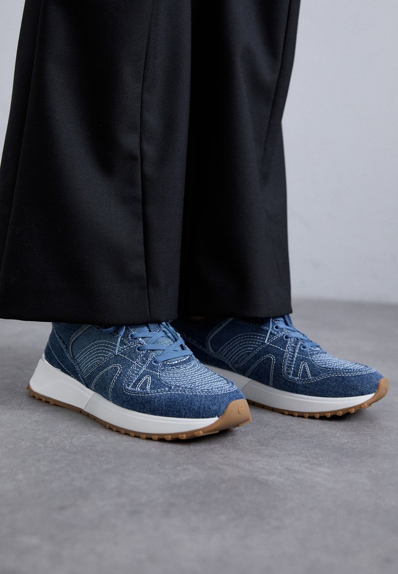 Blauwe denim sneakers met een gestructureerde bovenkant, witte tussenzolen en rubberen buitenzolen. De veters zijn in dezelfde blauwe kleur. Gecombineerd met losse zwarte broeken.