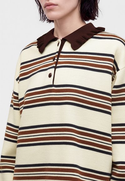 Personne portant un pull crème à rayures horizontales marron et noir et un col froncé brun foncé avec trois boutons.