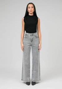 Femme avec de longs cheveux noirs portant un haut noir sans manches et un jean gris à taille haute et à jambes larges, se tenant devant un fond uni.