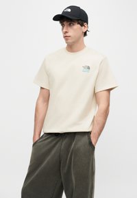 Άνδρας που φοράει μπεζ μπλουζάκι The North Face, μαύρο καπέλο και σκούρο χαλαρό παντελόνι, στέκεται με τα χέρια στις τσέπες, στραμμένος προς τα αριστερά.