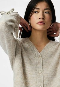 Young woman adjusting her long dark hair wearing silver hoop earrings and a beige knitted buttoned cardigan with tied sleeve cuffs.
