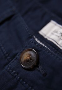 Primo piano di un tessuto blu navy con un grande bottone scuro e lucido a quattro fori, cucito sul tessuto, che mette in evidenza una texture raffinata.