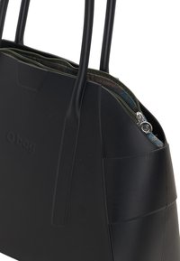 Zwarte handtas van zacht synthetisch materiaal, met ritssluiting, textuur interieur, twee stevige handgrepen en een opvallend logo aan de voorkant.