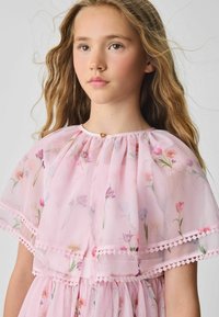Vaaleanpunainen kukkakuvioinen chiffon-mekko, jossa on röyhelökaulus, hennot pitsiyksityiskohdat ja hillitty kultainen solki kaulassa.