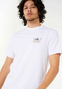 Hvid bomulds t-shirt med en grafisk front, der viser palmer og en solnedgang i orange og blå, mærket "RIP CURL THE SEARCH." Korte ærmer.