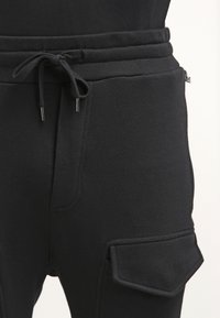 Svarta joggingsbyxor i mjukt tyg har ett elastiskt midjeband med snöre, sidofickor och en ficka på låret.