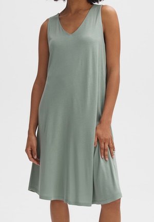 Vrouw die een mouwloze, licht saliegroene knielange jurk met V-hals draagt, staand tegen een effen lichte achtergrond.