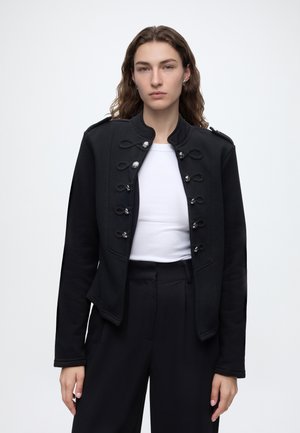 Jeune personne aux longs cheveux bouclés porte une veste noire style militaire par-dessus un haut blanc et un pantalon noir taille haute sur fond uni.