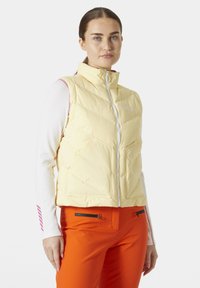 Světle žlutá zateplená vesta s vysokým límcem, předním zipem a dvěma zipovými kapsami; nošená přes bílou halenku s dlouhým rukávem a oranžovými kalhotami.