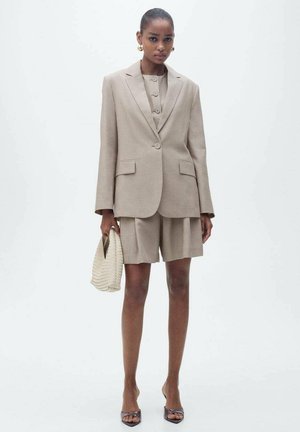 Blazer beige à rayures avec un short assorti, comportant un devant boutonné et deux poches. Le modèle tient un sac à main texturé de couleur claire.