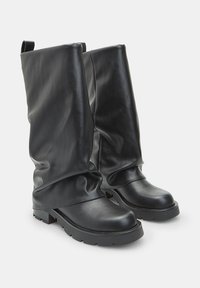 Alti stivali in pelle nera con canne larghe e spiegazzate e suole spesse in gomma su uno sfondo bianco plain.