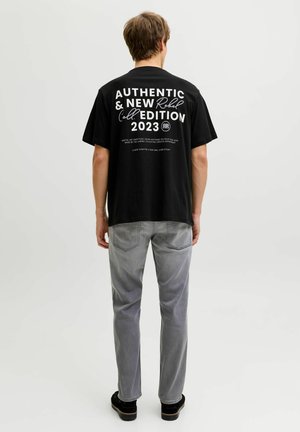 Uomo che indossa una maglietta nera con scritta bianca "Authentic & New Rebel Edition 2023" sulla schiena, jeans grigi, scarpe nere, in piedi di spalle.