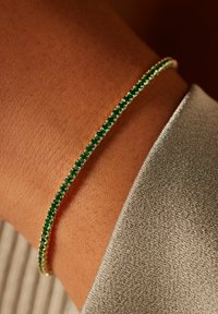 Emerald green bracelet with small gemstones set in a thin gold band, worn on tanned skin, against a textured silver fabric background.