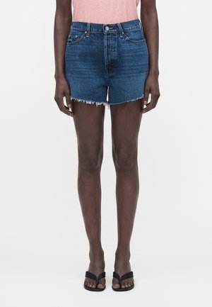 Person iført blå denim kortbukser, lyserød skjorte og sorte flip-flops står foran en ensfarvet hvid baggrund.