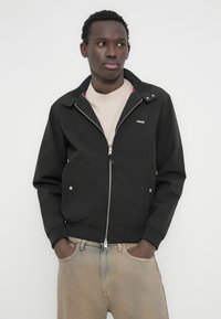 Νέος άνδρας με σκουρόχρωμο δέρμα, φορώντας μαύρο τζάκετ με φερμουάρ, μπεζ μπλουζάκι και ξεθωριασμένο ανοιχτό μπλε τζιν, στέκεται με τα χέρια στις τσέπες.