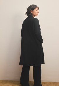 Lang, sort, ribbet cardigan med en lige silhuet, parret med løse, sorte bukser. Stoffet har en tekstureret overflade og sidelommer.