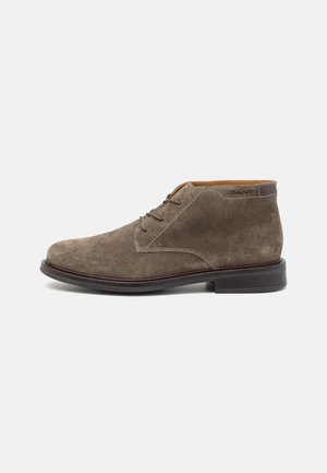 Braune Wildleder-Stiefeletten mit runder Zehenpartie, ausgestattet mit Schnürsenkeln, kontrastierenden Nähten und einer dunklen Gummisohle. Geprägtes Logo an der Seite.