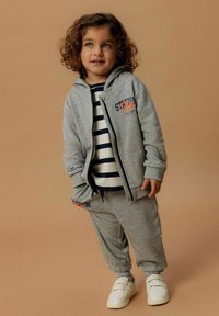 Grijze zip-up hoodie met oranje logo, gestreept shirt eronder, bijpassende grijze joggingbroek en witte sneakers met klittenbandsluitingen.