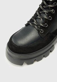 Bottines noires avec une tige en cuir texturé, des accents en daim et une semelle en caoutchouc épaisse avec une rainure marquée. Les lacets sont noirs avec des rayures blanches.
