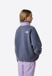 Flis pulover v mehki sivkini barvi z teksturiranim sivim telesom, visokim ovratnikom in vezenim logotipom na hrbtu. Ohlapen kroj in padajoča ramena.