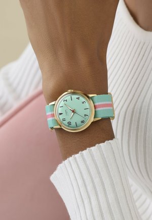 Karóra menta zöld számlappal, arany tokban és pasztell zöld, rózsaszín és fehér csíkozású szövet szíjjal, egy személy csuklóján, aki fehér bordázott ujjú felsőt visel.