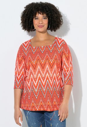 Kiharatukkainen nainen, jolla on oranssi siksak-kuvioinen pusero, jossa on kolme neljännestä hihaa, ja siniset maalitahroilla pilkulliset farkut, seisoo valkoista taustaa vasten.