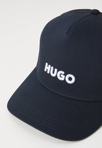 Mørkeblå caps laget av stoff med flat skjerm. Har hvit brodert "HUGO" logo på forsiden. Teksturert mesh-panel på baksiden.