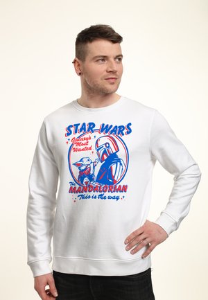 Mand iført hvid sweatshirt med rød og blå Star Wars Mandalorian grafik og tekst, kigger til siden med den ene hånd på hoften.