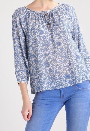 Blouse à manches longues à motif floral bleu et blanc avec un col à nouer, associée à un jean bleu clair.