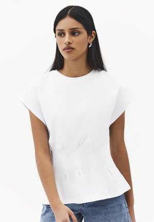 Junge Frau mit glatten schwarzen Haaren, die ein weißes strukturiertes Oberteil und blaue Jeans trägt und leicht seitlich vor einem weißen Hintergrund schaut.