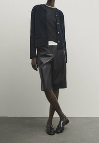 Marineblauer strukturierten Cardigan mit goldenen Knöpfen, über einem schwarzen Oberteil getragen, kombiniert mit schwarzer Leder-Shorts und Loafers. Schlichte, klare Linien.