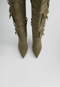 Olivgröna, knähöga stiletto stövlar med en texturerad finish, spetsiga tår och justerbara remmar med guldfärgad hårdvara.